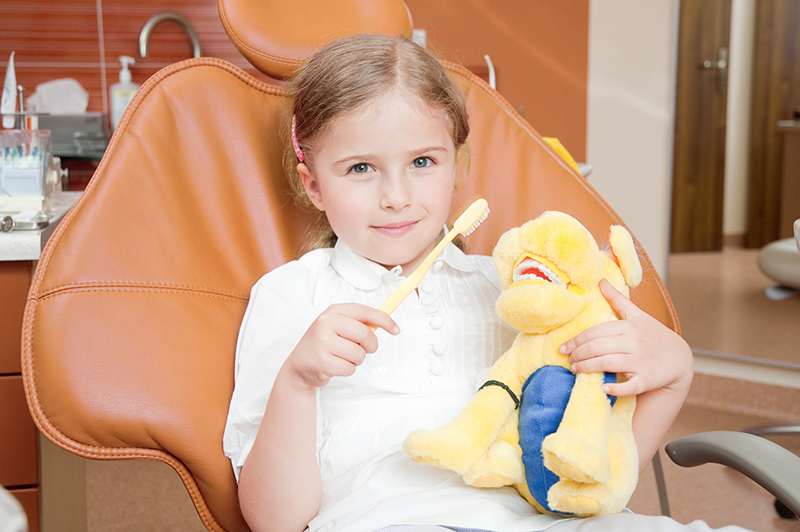 young girl in leather operatory chair with toothless smile holds a yellow toothbrush and stuffed animal