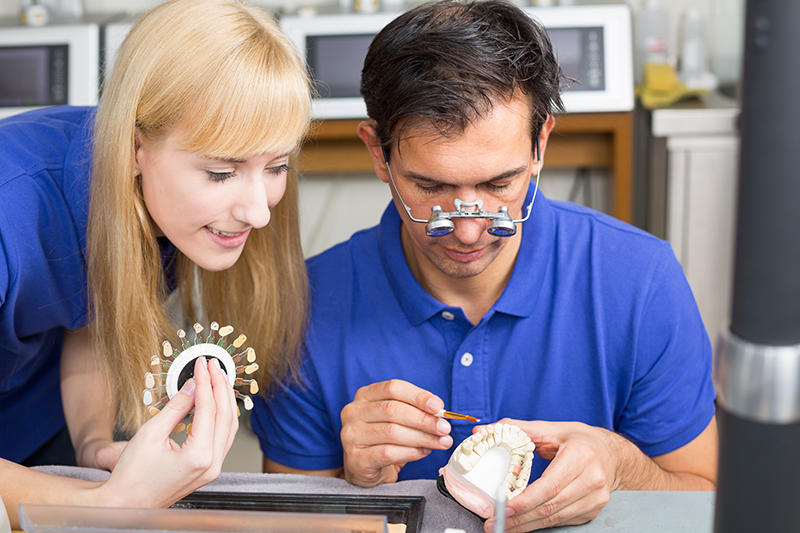 male dentist looking at example of teeth with magnifying glasses as female assistant looks on