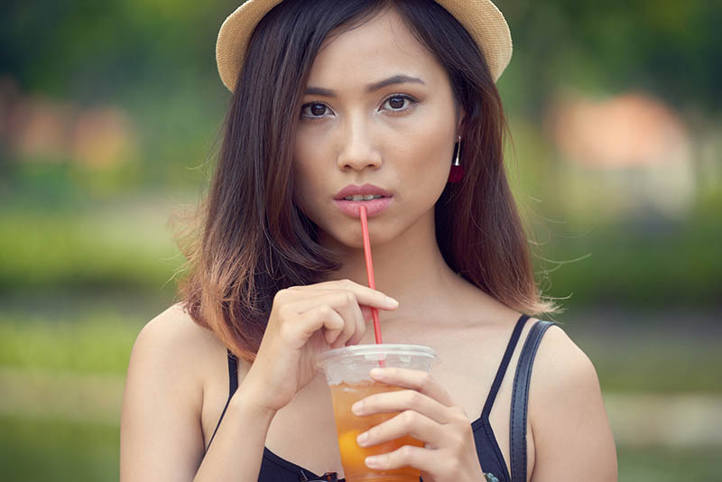 asian woman with a hat in a park drinking tea in a plastic cup from a straw