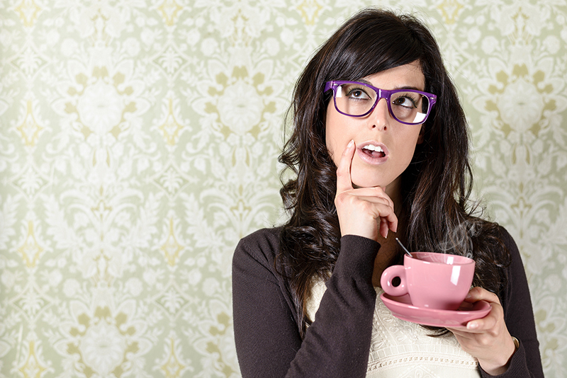 brunette woman holding her chin wearing purple reading glasses and holding a pink tea cup with a spoon
