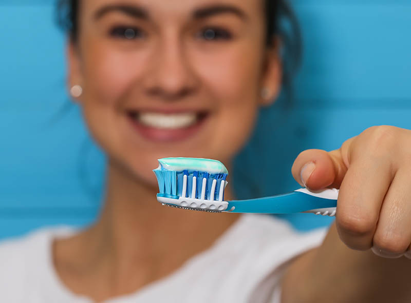 woman blurred holding out an in focus blue toothbrush with toothpaste on the bristles